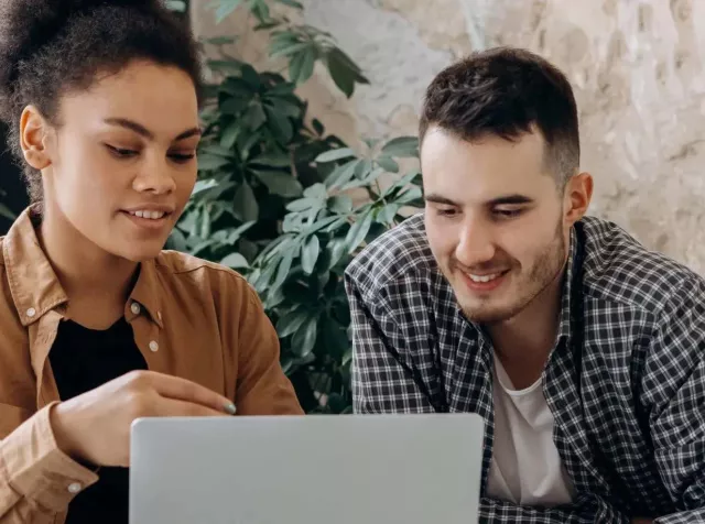 Ein Mann und eine Frau sitzen zusammen und schauen auf einen Laptopbildschirm. Beide wirken konzentriert und lächeln – die Szene zeigt Zusammenarbeit und gemeinsames Arbeiten am Computer.