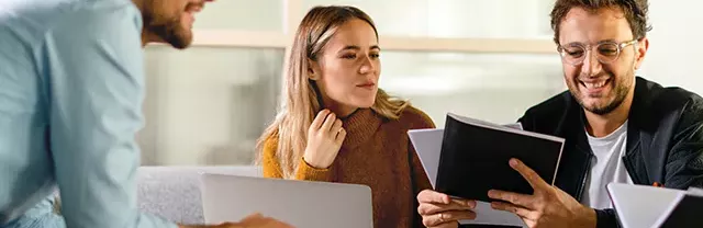 Zwei Männer und eine Frau sitzen zusammen auf einem Sofa und arbeiten gemeinsam mit Laptops und Dokumenten. Sie unterhalten sich und lachen, die Szene zeigt Zusammenarbeit und Teamwork.